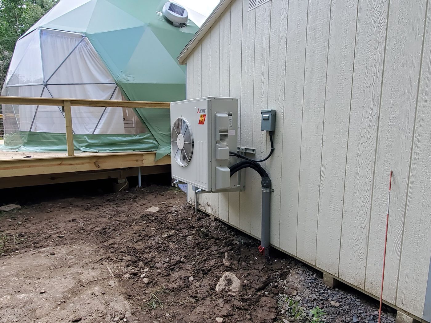 Mitsubishi Electric Hyper-Heating outdoor unit on wall brackets on the side of a shed next to the green geodesic dome