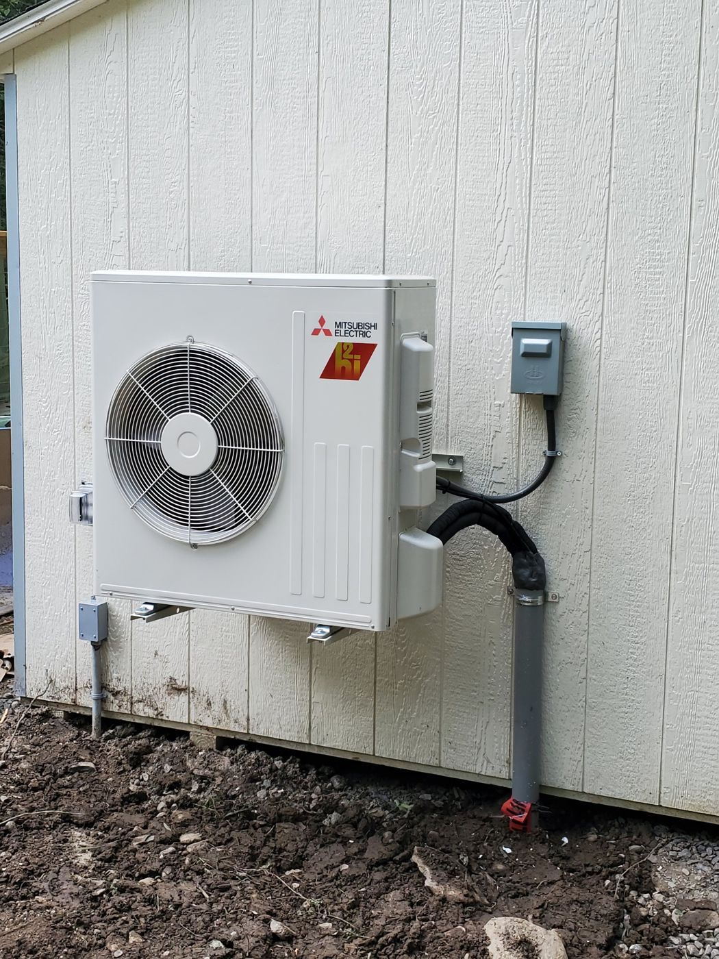 Close-up of a Mitsubishi Electric H2i Hyper-Heating outdoor unit with an electrical disconnect and the insulated line set running down a gray conduit into the ground