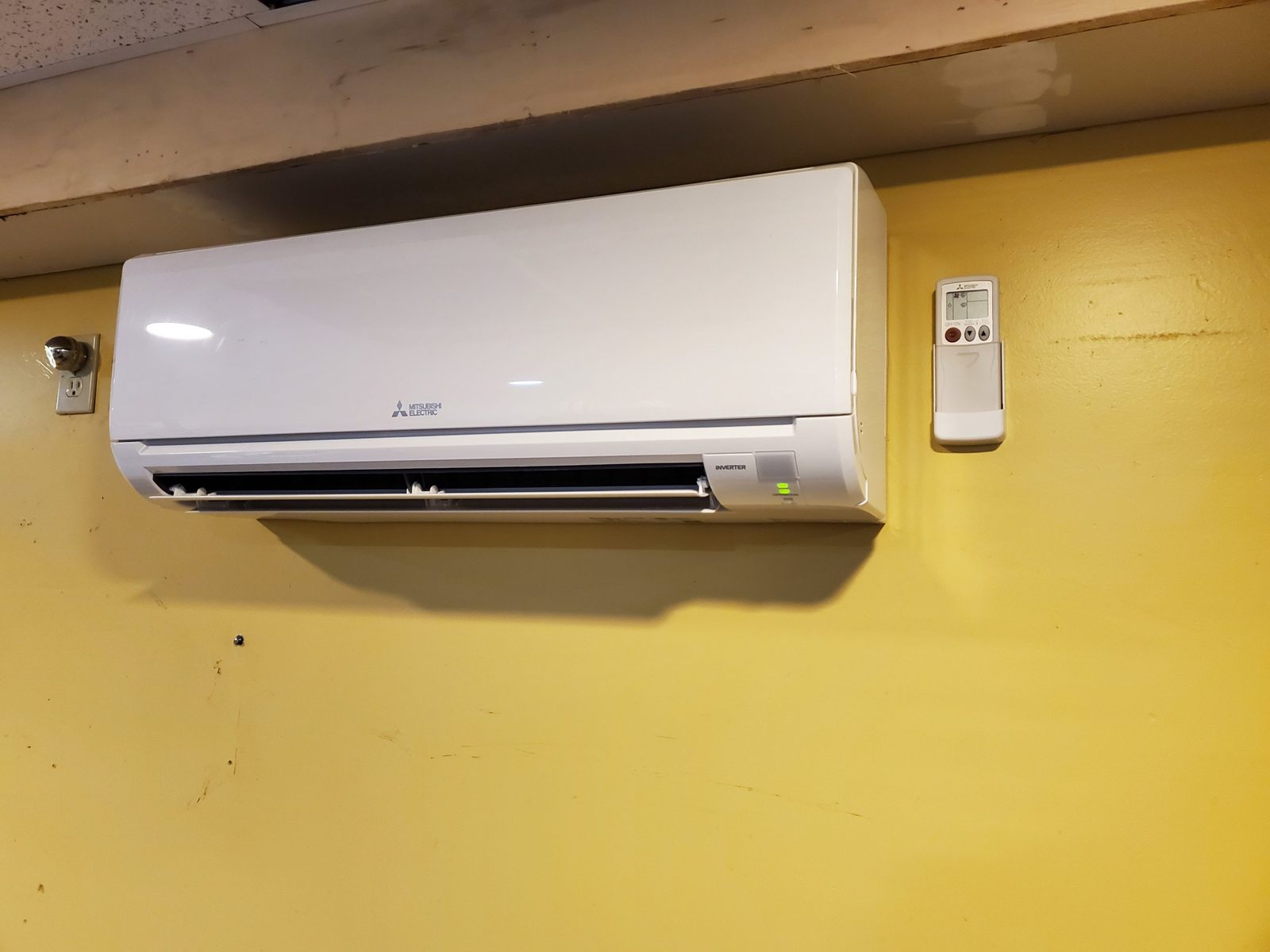 Mitsubishi Electric ductless wall unit on a yellow wall under a beam, with its remote in a holder beside it