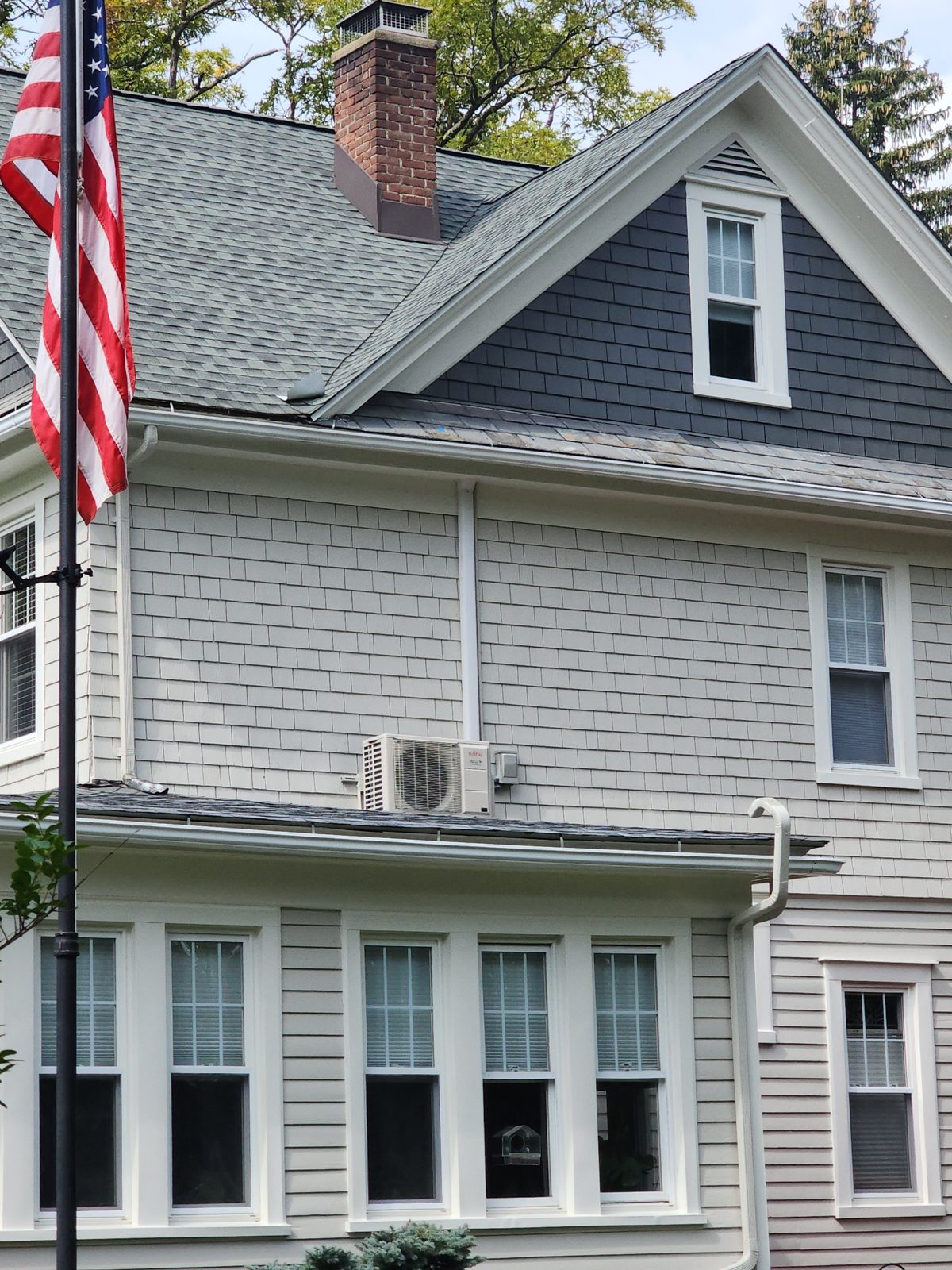 Fujitsu heat pump outdoor unit set on a porch roof against gray shingle siding, with a white line-set cover running up the wall beside the downspout